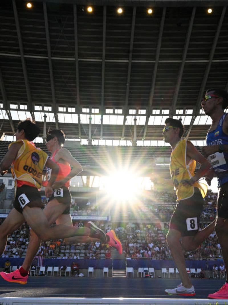 two vision impaired runners and their guides running on the track in front of spectators 